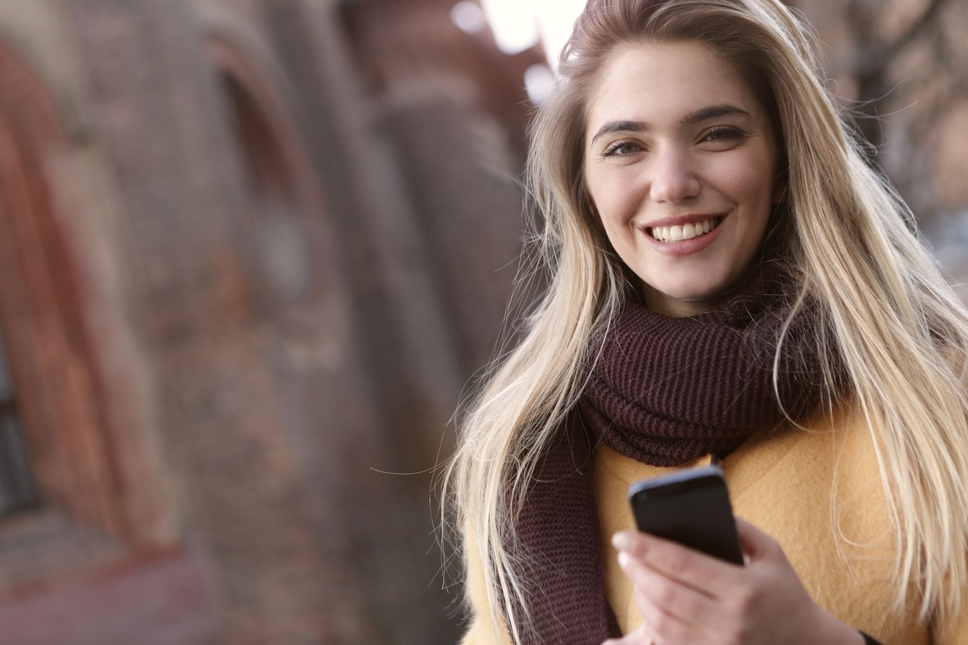 Woman with long light brown hair wearing mustard yellow jacket and dark turtleneck, smiling while holding smartphone, standing outside brick building.