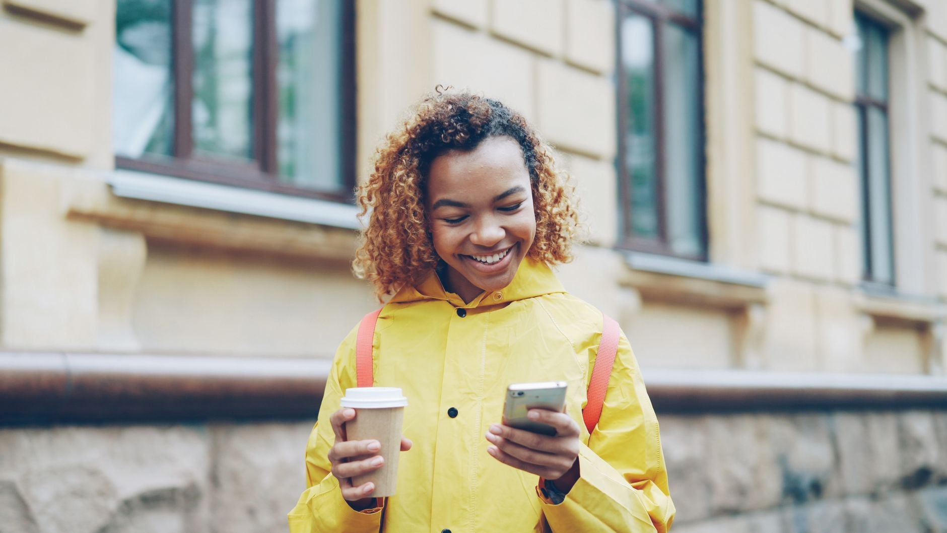 Young woman with reddish-brown curly hair wearing bright yellow jacket with red backpack straps, holding coffee cup and smartphone outdoors in front of building.